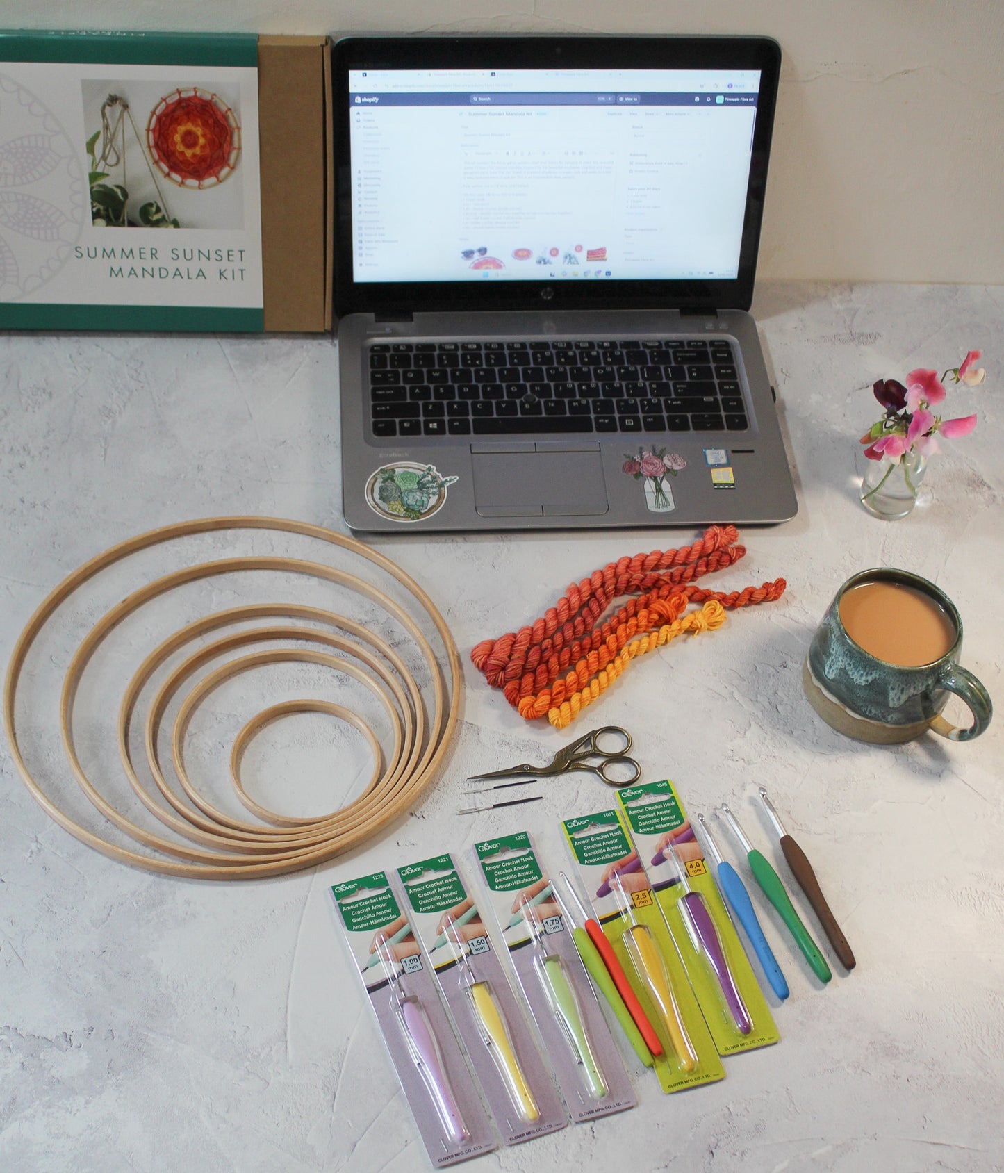 dClover Armor crochet hooks, wooden rings for crochet, macrame or other craft, needles, scissors and yarns arrange around an open laptop, a cup of tea and a small vase of sweetpeas