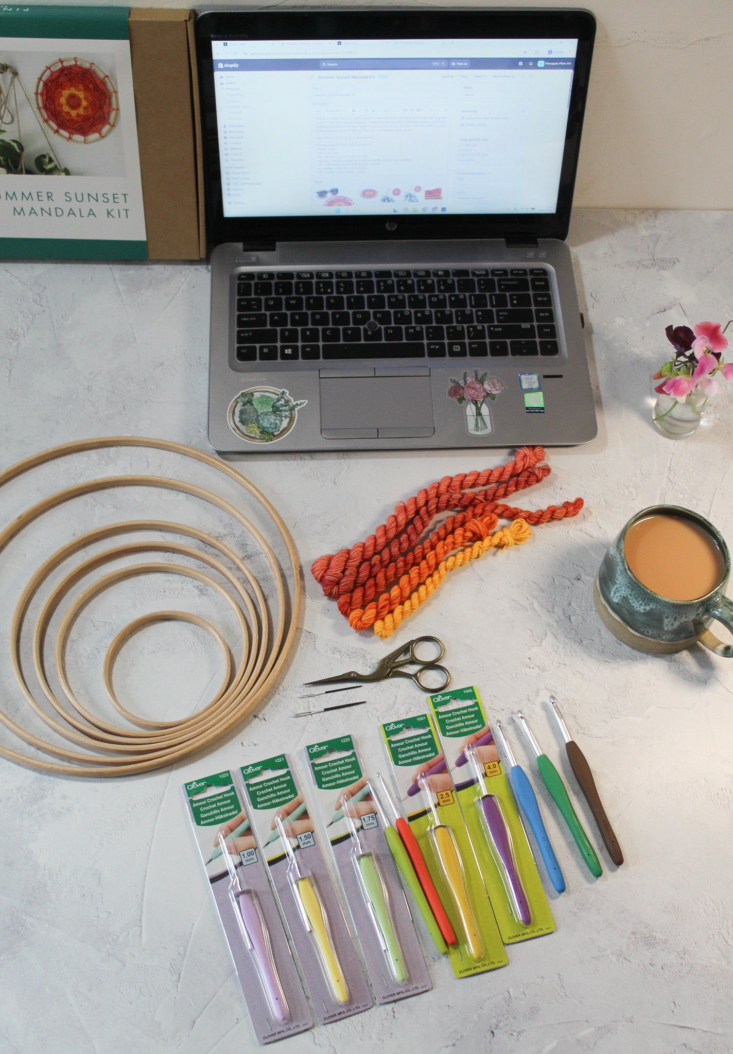 dClover Armor crochet hooks, wooden rings for crochet, macrame or other craft, needles, scissors and yarns arrange around an open laptop, a cup of tea and a small vase of sweetpeas