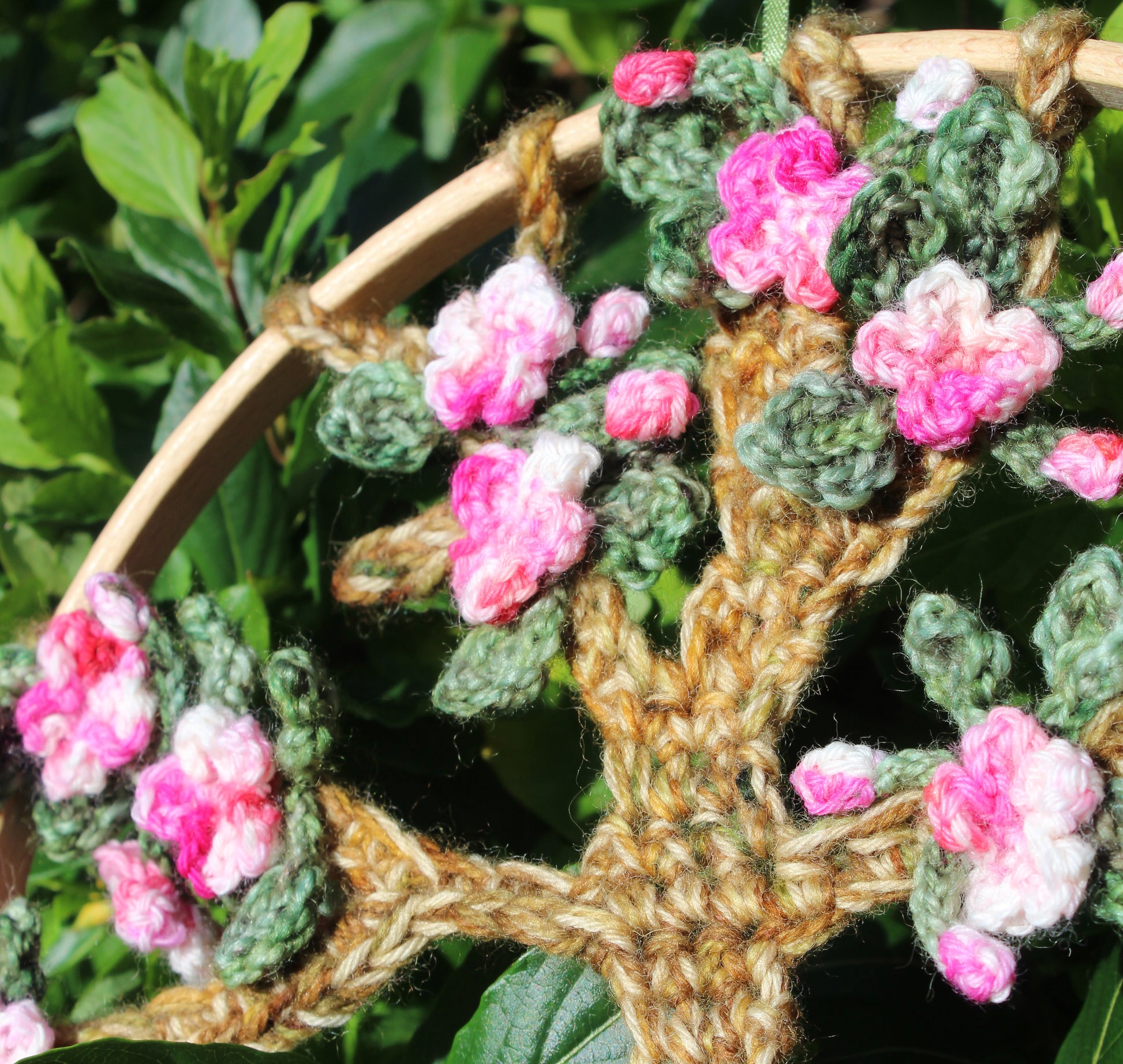 Crocheted floral wreath with pink and green flowers on a wooden hoop against a natural background.