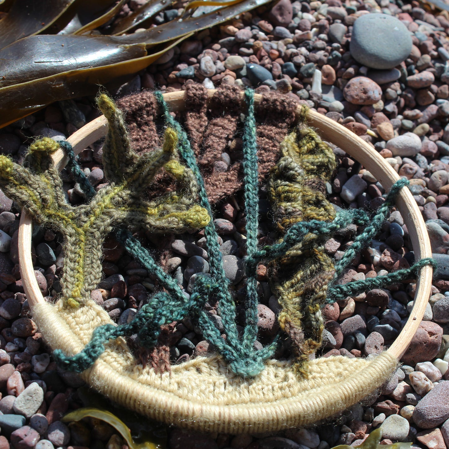 A crochet mandala you can make of British seaweeds, shown on a pebbly beach with real seaweed.